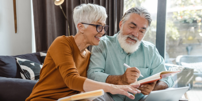 An older couple smiles while working together on a laptop and taking notes.